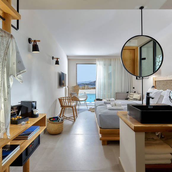 One bedroom suite with aa double bed, open vanity area, clothes hanger, wooden furniture, and a balcony with a pool and sea view